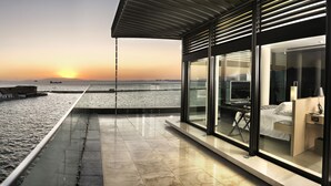 Quarto de hotel moderno com uma ampla janela de vidro que oferece vista para a orla e para o pôr do sol.
