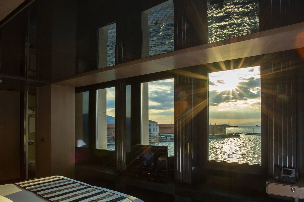 A hotel room with a large window overlooking a body of water, a bed with a striped bedspread, and a desk with a telephone.