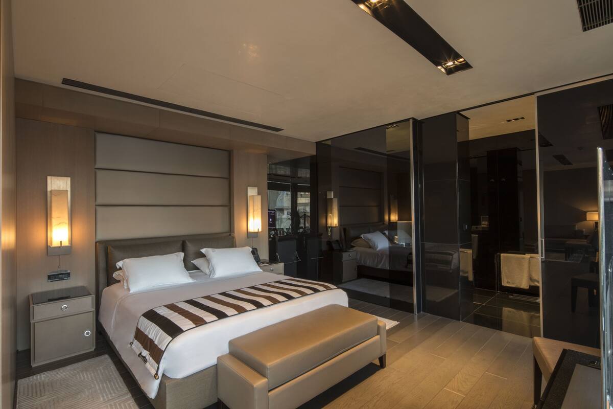 A modern bedroom with a large bed, bedside tables, a bench, and a glass partition.