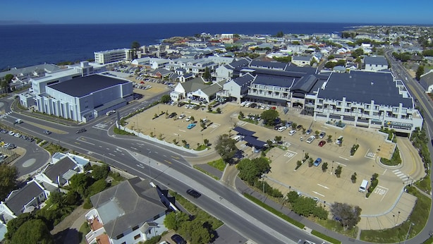 An aerial view of a coastal town with a large parking lot, a prominent building with a flat roof, and a road leading to the sea. - Whale Coast All Suite Hotel (Hermanus)