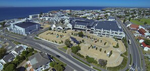Une vue aérienne d’une ville côtière, avec un grand parking, un bâtiment imposant doté d’un toit plat et une route menant à la mer.