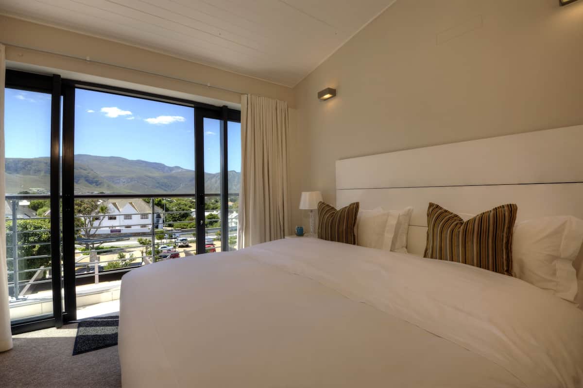 A large bed with white linens, a nightstand with a lamp, and a view of mountains through large windows.