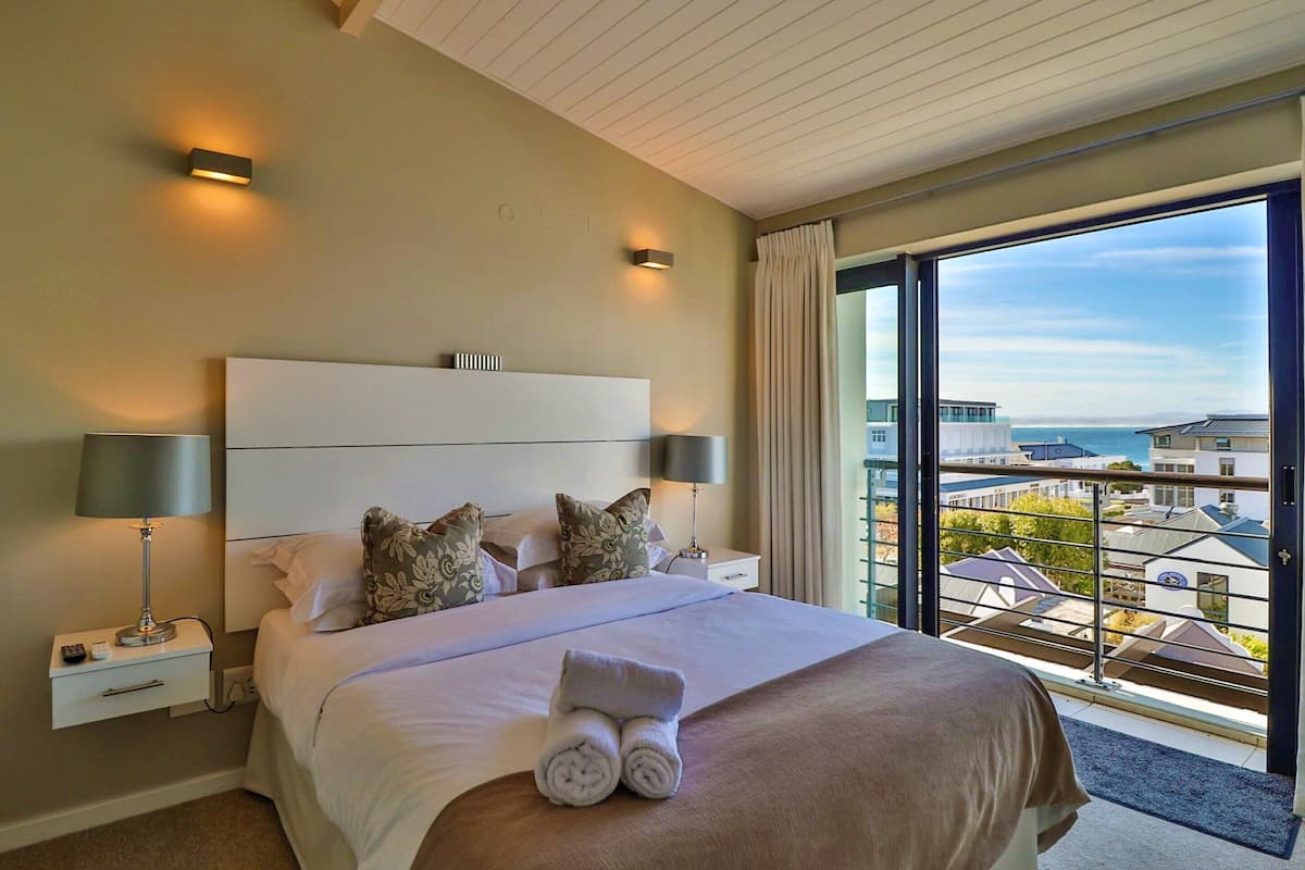A hotel room with a large bed, two bedside lamps, a nightstand, and a view of buildings through a large window.