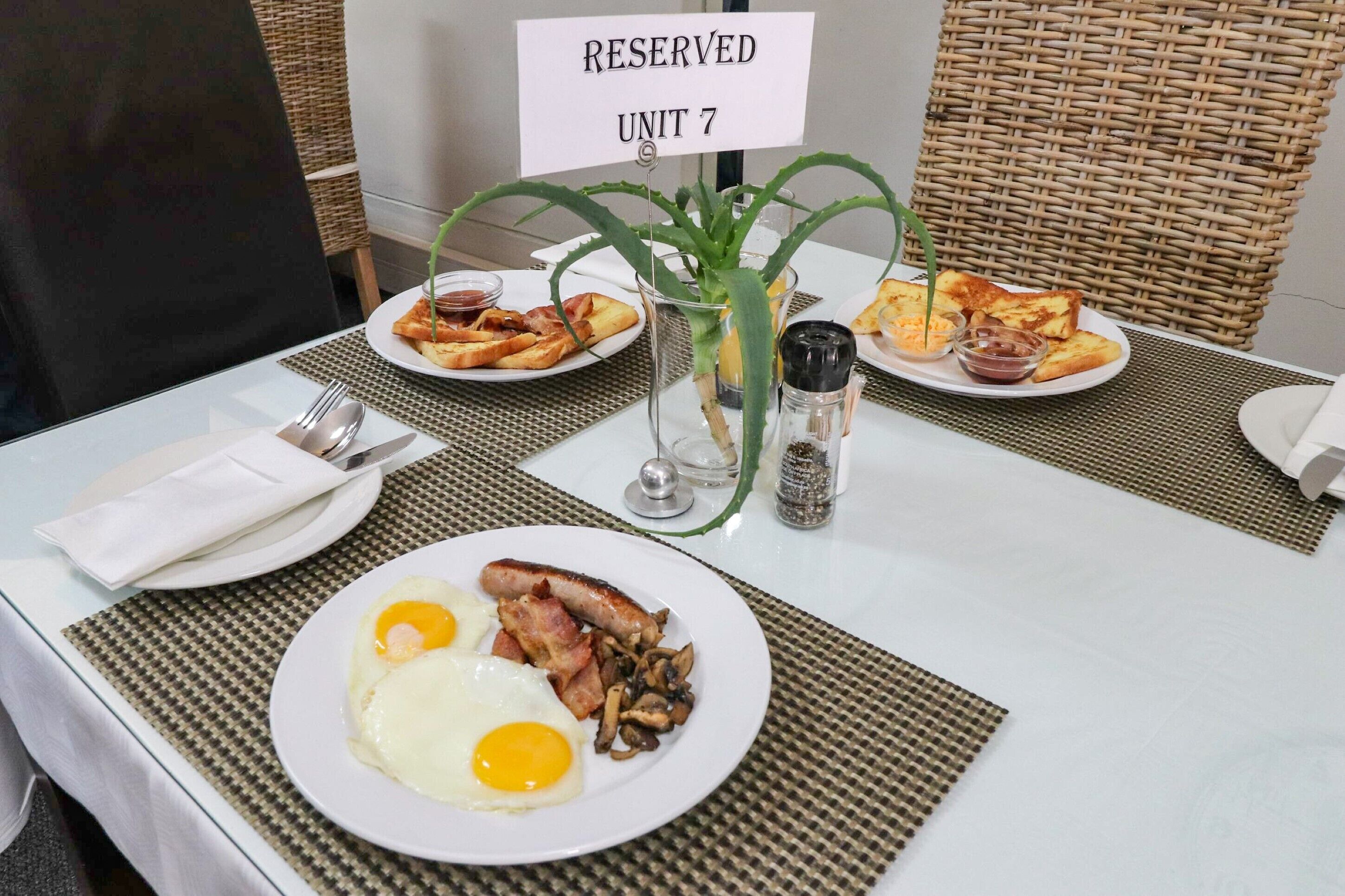 Una mesa de desayuno con platos de huevos, tocino y champiñones, un pequeño jarrón con plantas verdes, y un letrero que indica 'RESERVED UNIT 7'.