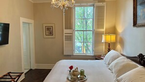 Ein Schlafzimmer mit einem großen Bett, einem Chandelier, einem Fenster mit Fensterläden, einem Nachttisch mit Lampe und einem gerahmten Bild an der Wand.