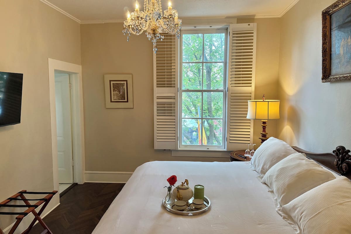 A bedroom with a large bed, a chandelier, a window with shutters, a bedside table with a lamp, and a framed picture on the wall.
