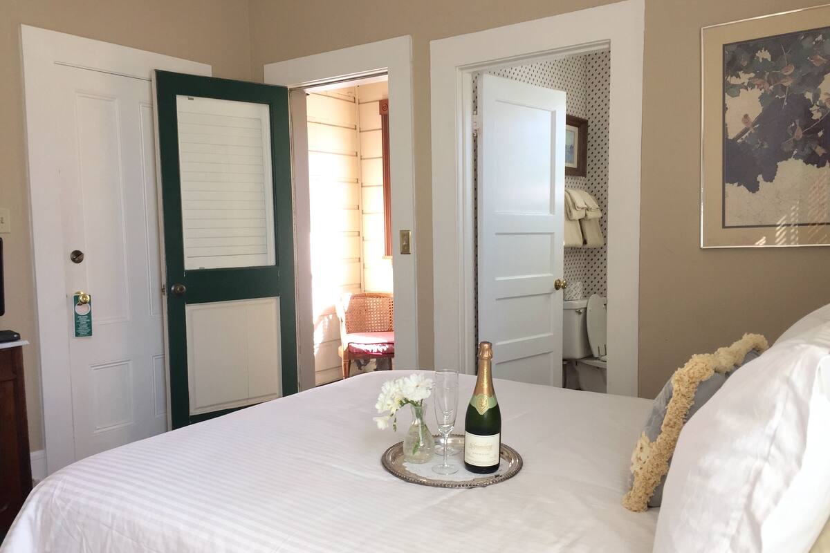 A hotel room with a bed, a tray with a bottle and glasses, and a door with a green frame.