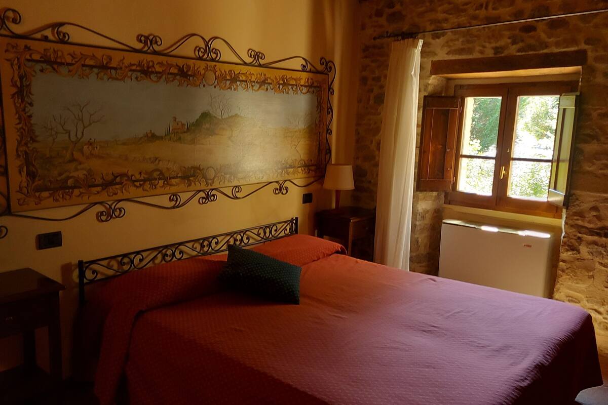 Une chambre à coucher avec un lit, une table de chevet, une lampe, une fenêtre avec des rideaux et un mur en pierre.