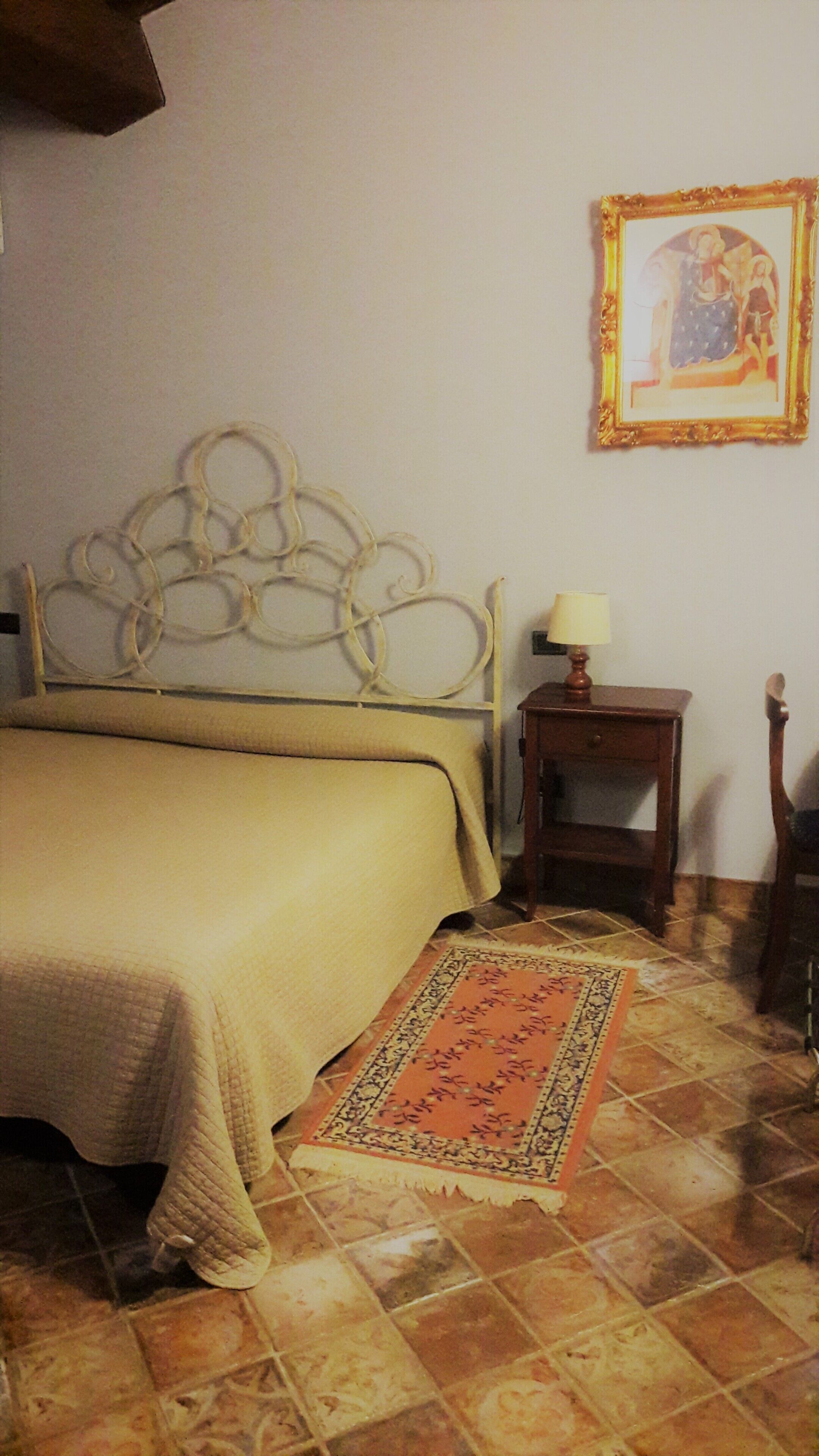Un dormitorio con cama, mesita de noche, lámpara, cuadro en la pared y alfombra estampada.