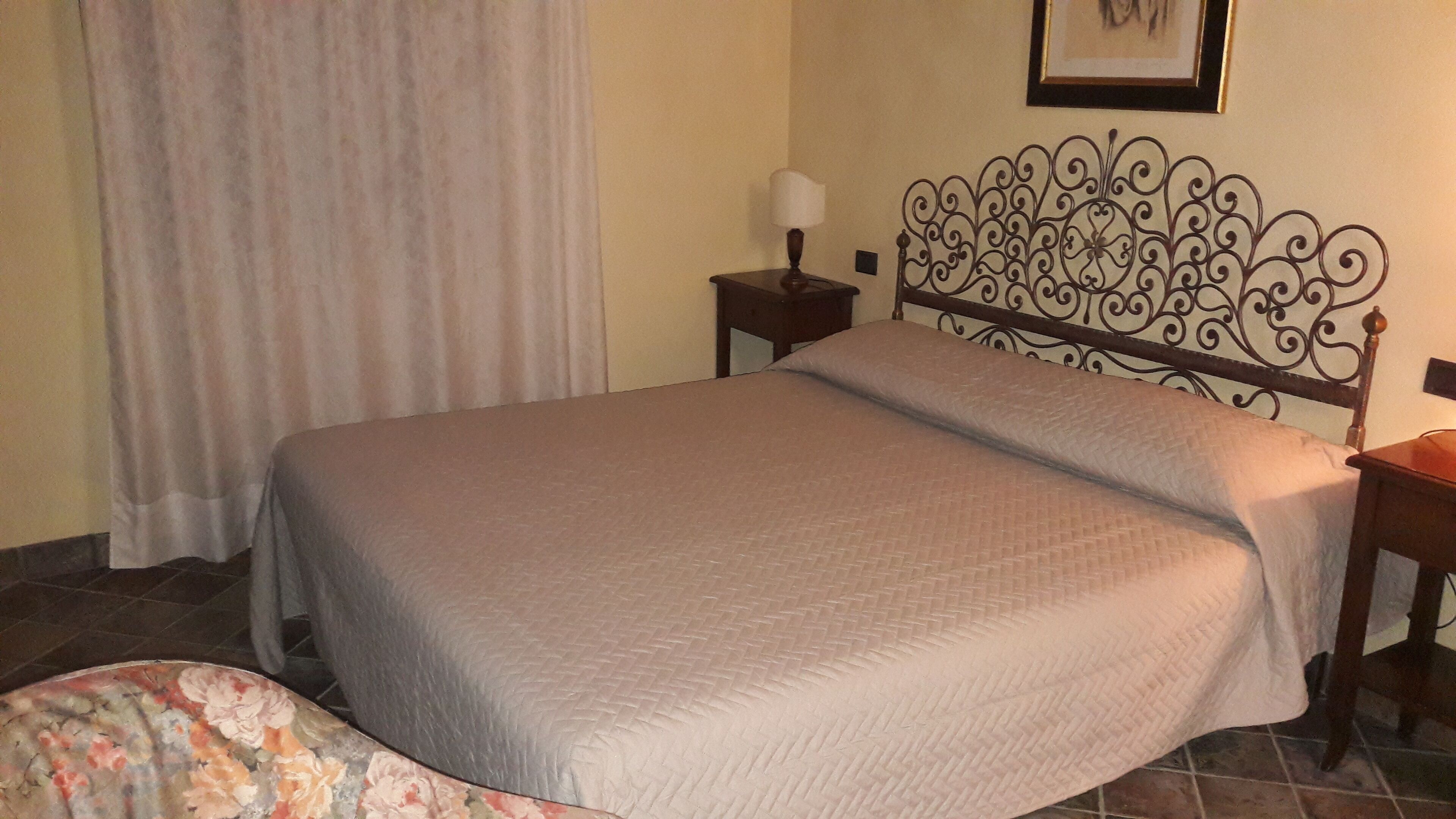 Una cama bien hecha con cabecera, una mesita de noche con lámpara y una cortina en la ventana.