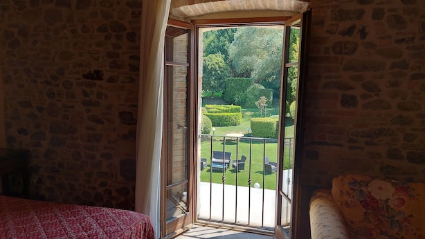 Une pièce avec un mur en pierre, un lit recouvert d’une courtepointe à motifs rouges, et une vue sur un jardin bien entretenu avec une pelouse, des haies et du mobilier d’extérieur.