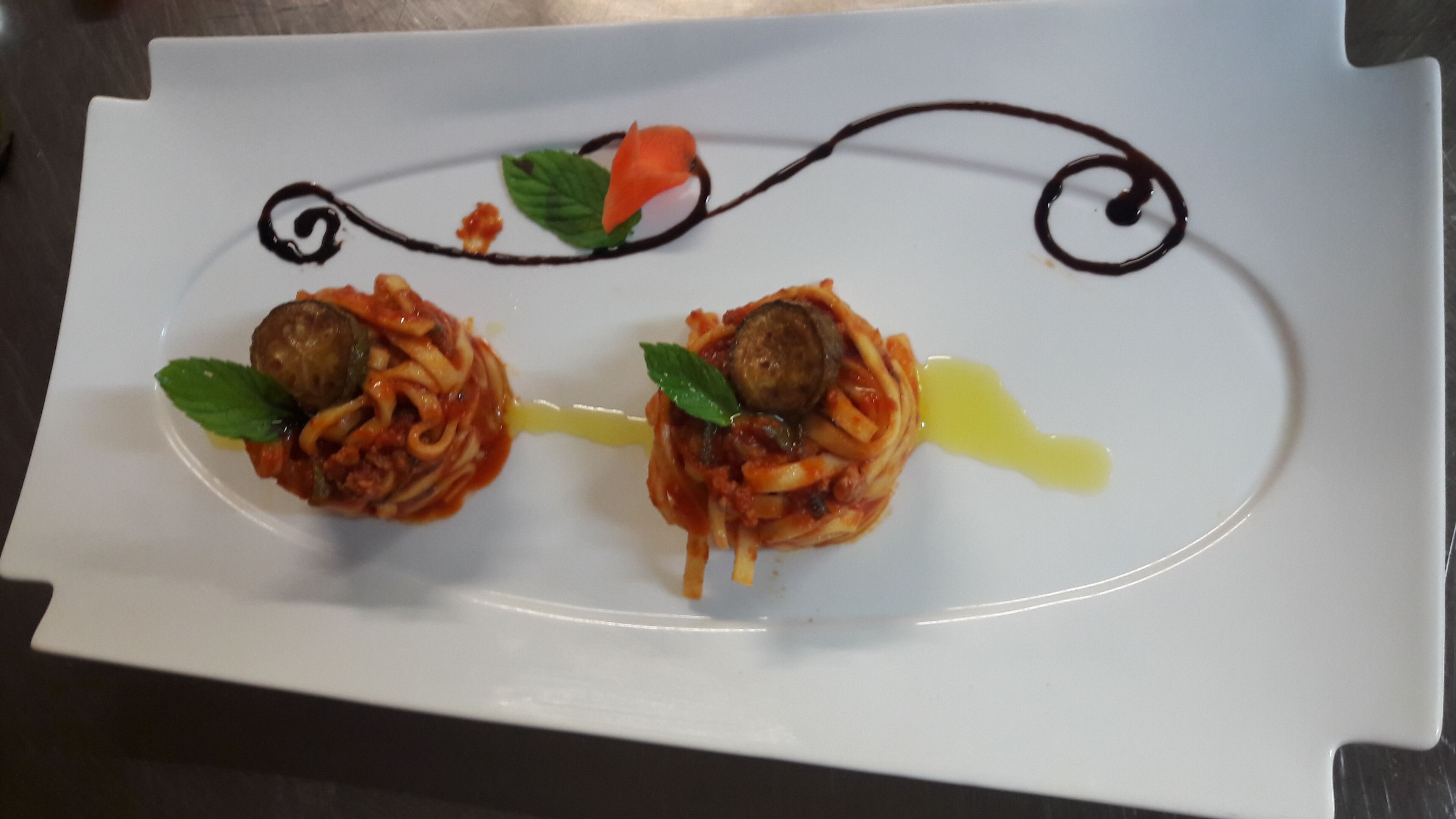 Un plato blanco con dos platos de pasta, cada uno cubierto con una albóndiga y una rodaja de tomate, adornado con una hoja de albahaca.