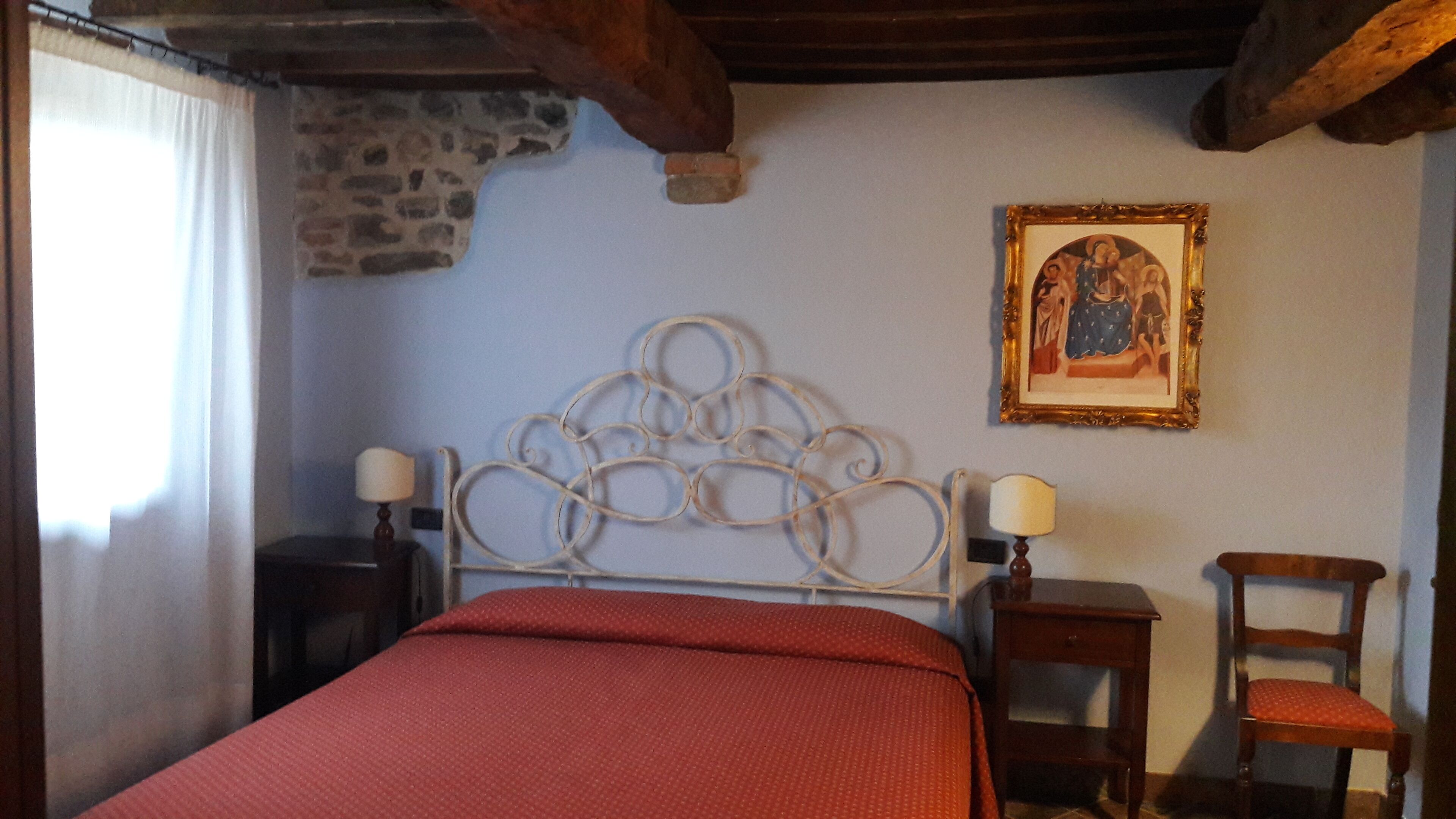 A bedroom with a stone wall, a metal bed frame, a red bedspread, two bedside tables with lamps, a wooden chair, and a framed picture on the wall.