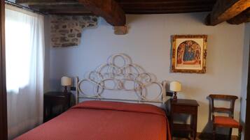 A bedroom with a stone wall, a metal bed frame, a red bedspread, two bedside tables with lamps, a wooden chair, and a framed picture on the wall.