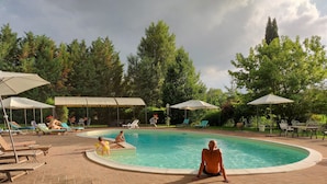 Un espace piscine agrémenté de chaises longues et de parasols, entouré d’arbres.