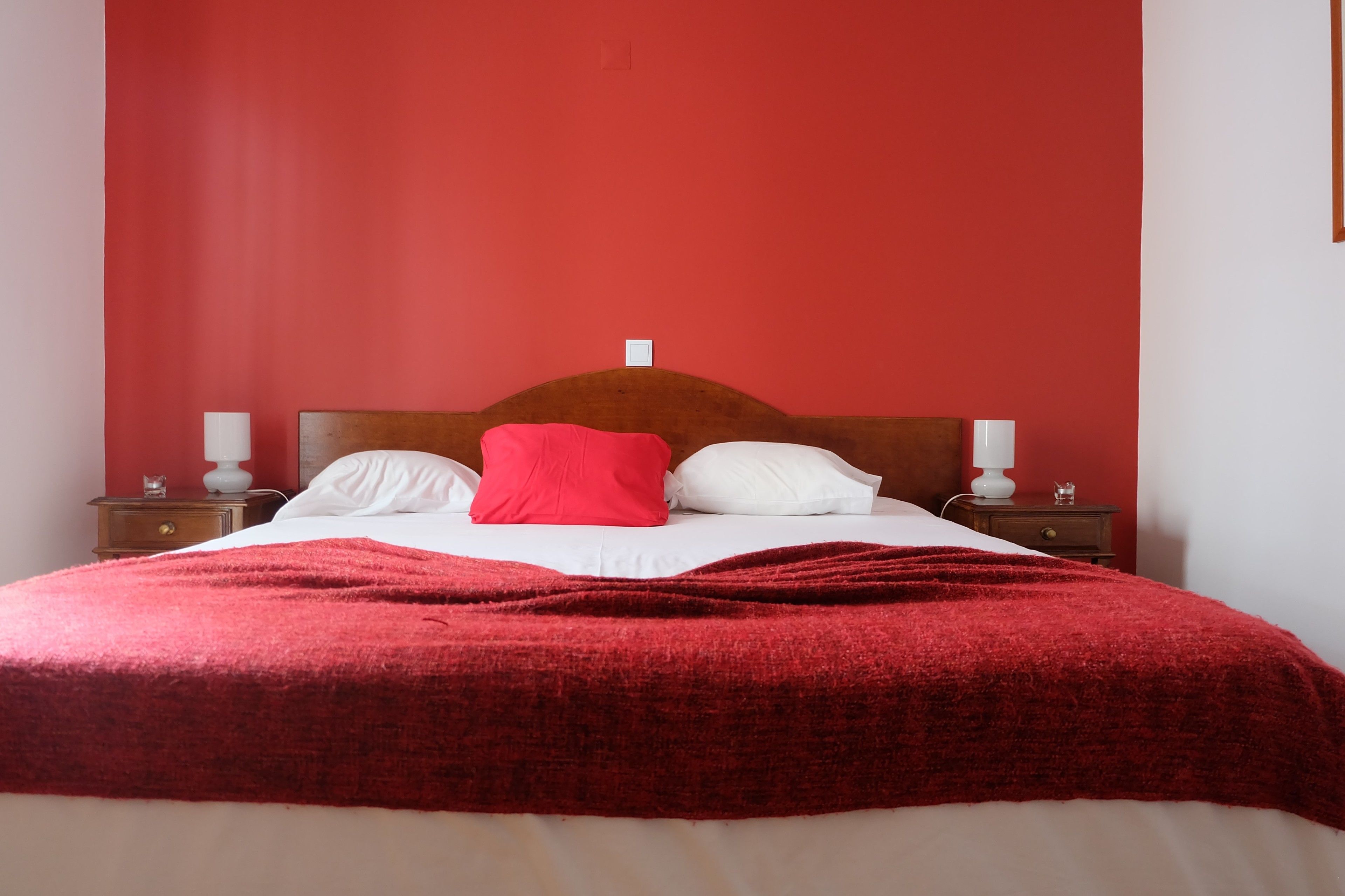 A bed with a red blanket, white pillows, and a wooden headboard. There are two bedside tables with lamps and a small decorative item on each.