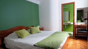 A bedroom with a bed, green walls, a wooden floor, a desk with a chair, and a bathroom with a green-tiled sink.