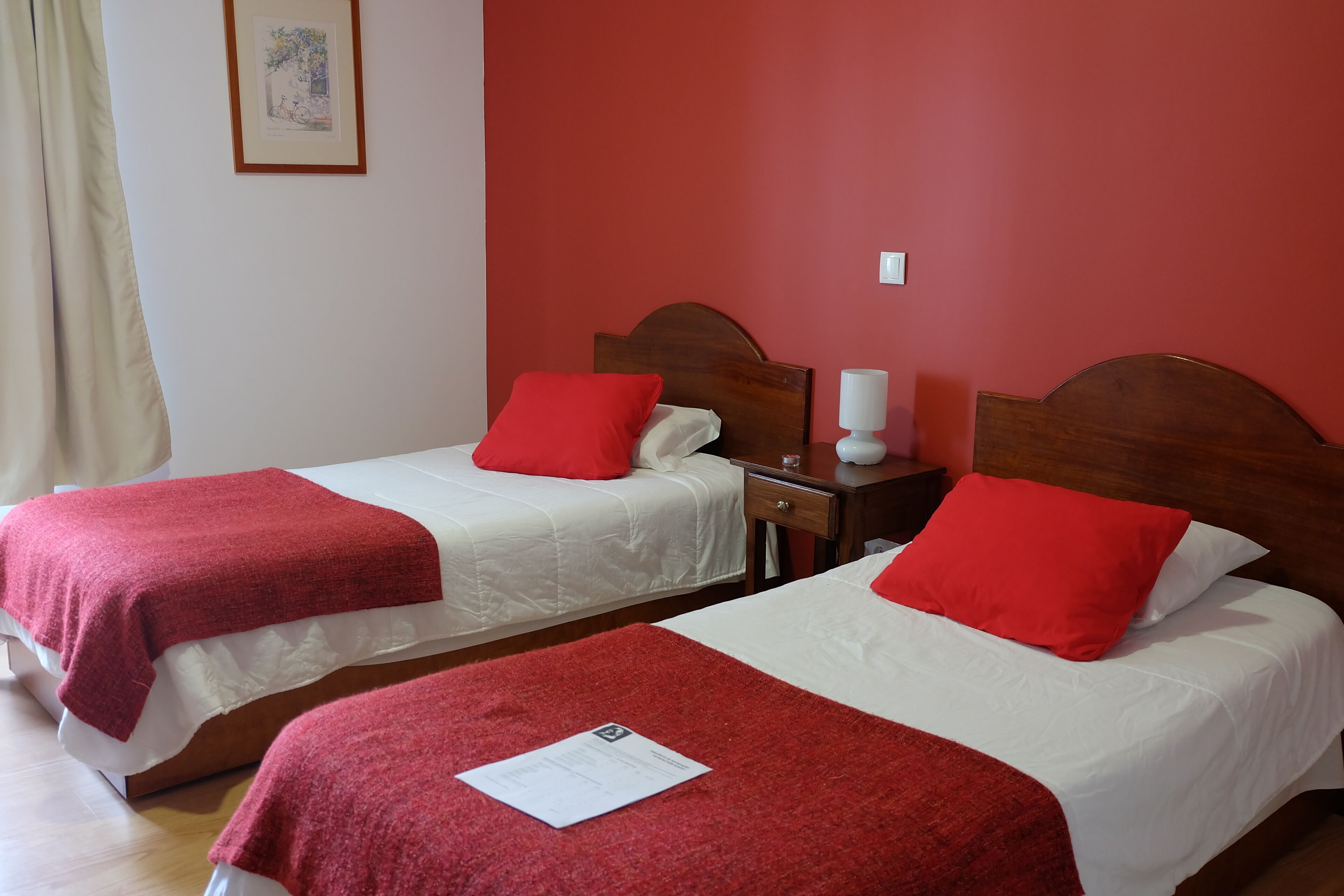 Deux lits simples avec une literie rouge et blanche, une tête de lit en bois, une table de chevet avec une lampe et un tableau encadré au mur.