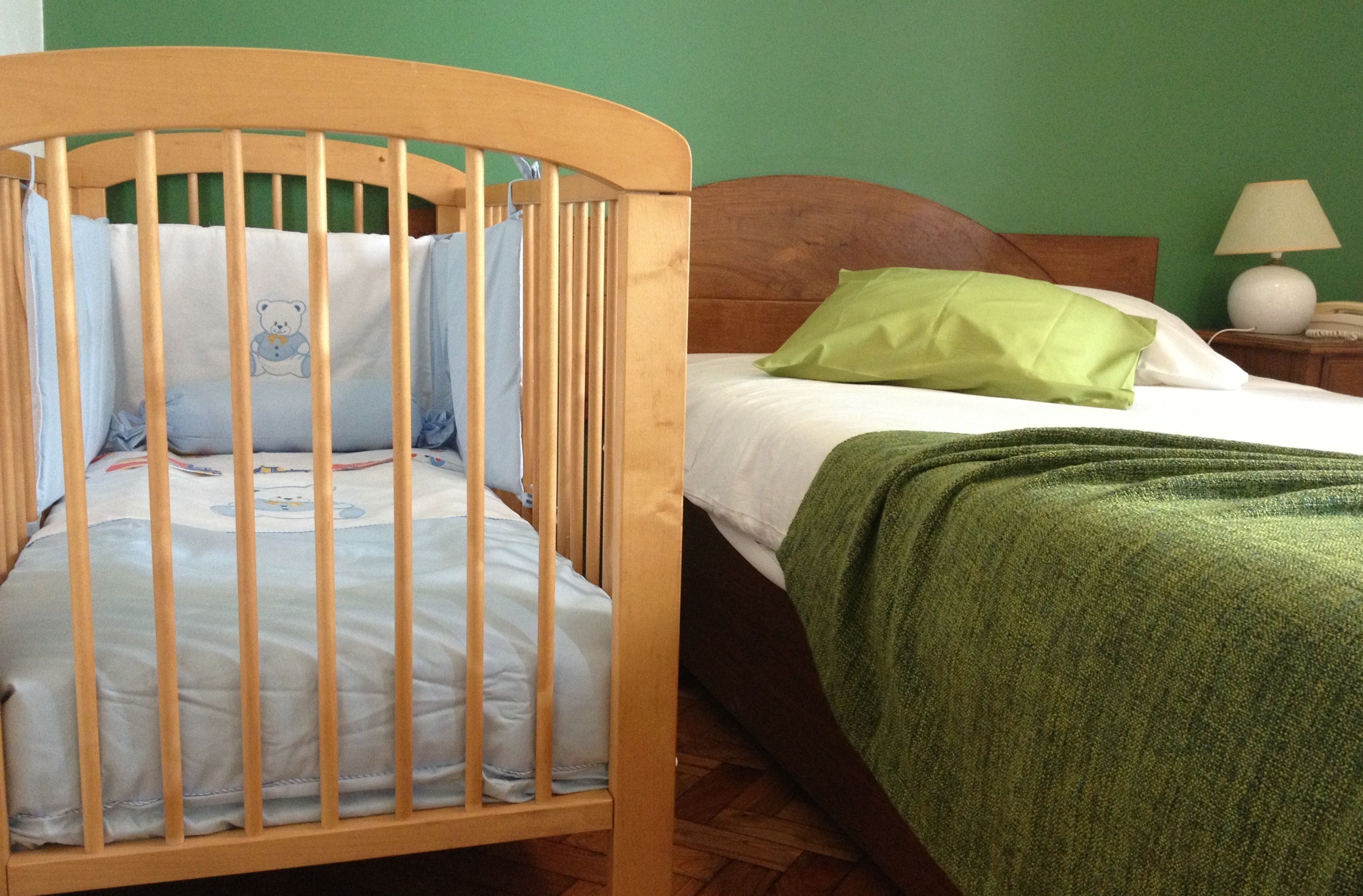 Quarto com berço de madeira, cama com manta verde e mesa de cabeceira com abajur.
