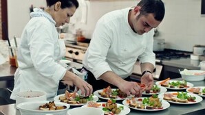 Dos cocineros preparando comida en una cocina con electrodomésticos de acero inoxidable y encimeras blancas.