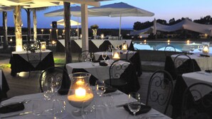 Un área de comedor al aire libre con mesas preparadas para dos, velas y una piscina al fondo.