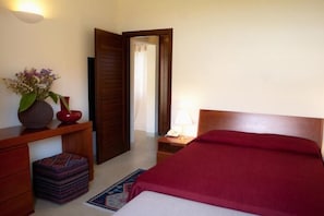 Une chambre avec un lit en bois, une table de chevet avec une lampe, un petit tapis et une étagère murale ornée d’un vase de fleurs.
