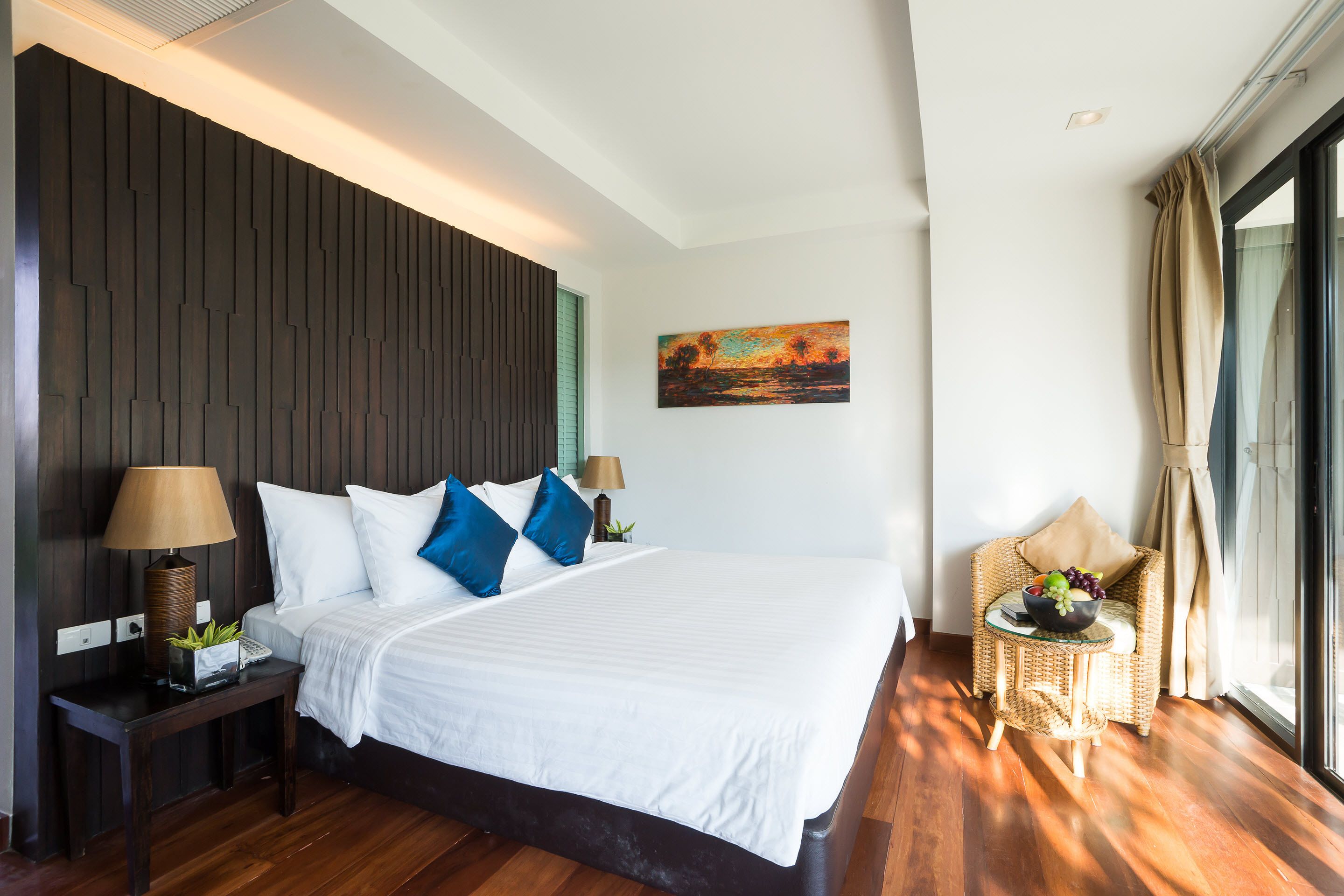 A modern hotel room with a large bed, two bedside tables with lamps, a small side table with a bowl of fruit, a wicker chair, and a painting on the wall.