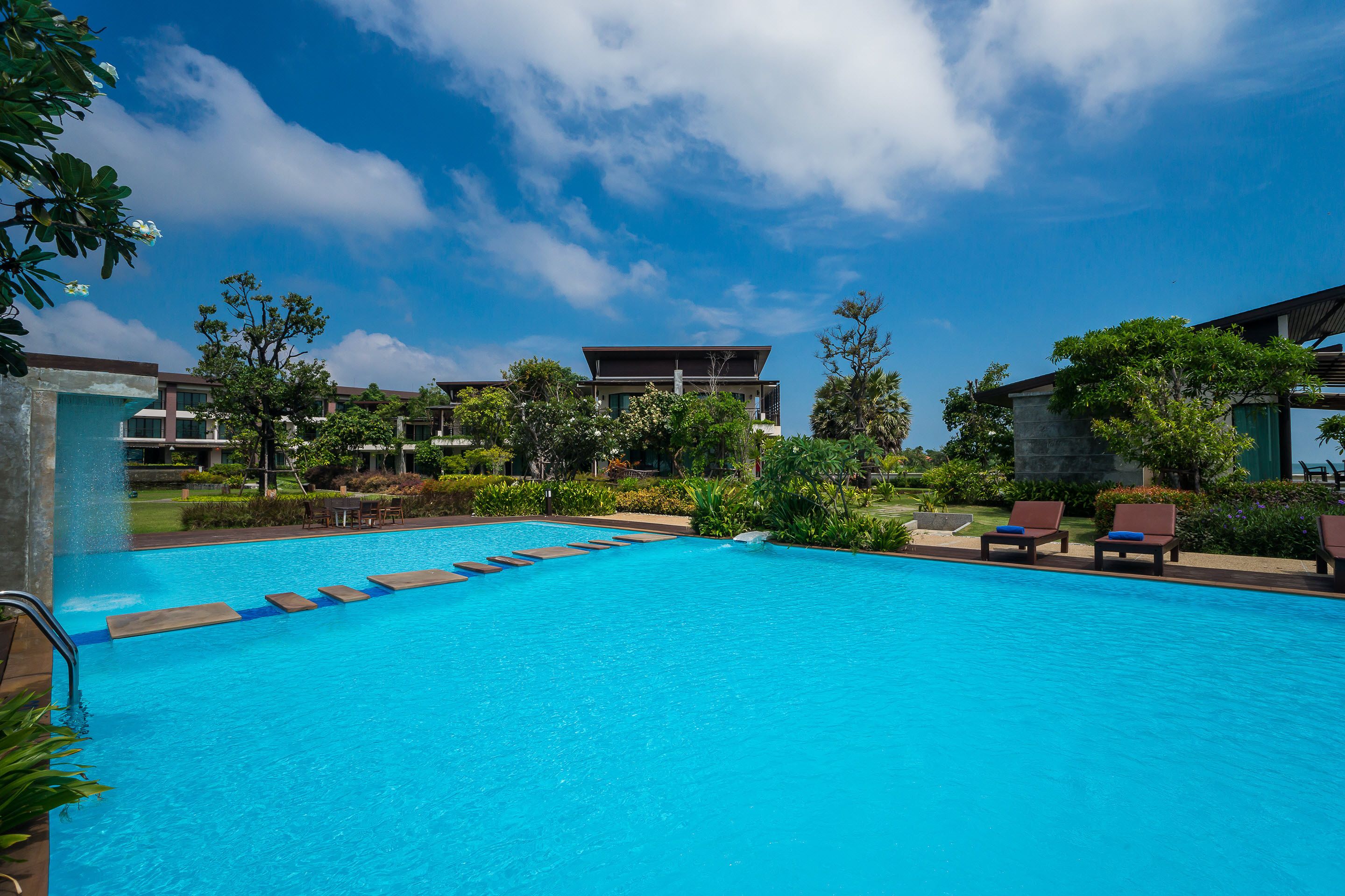 Una piscina con agua azul cristalina, rodeada de sillas de descanso y follaje.