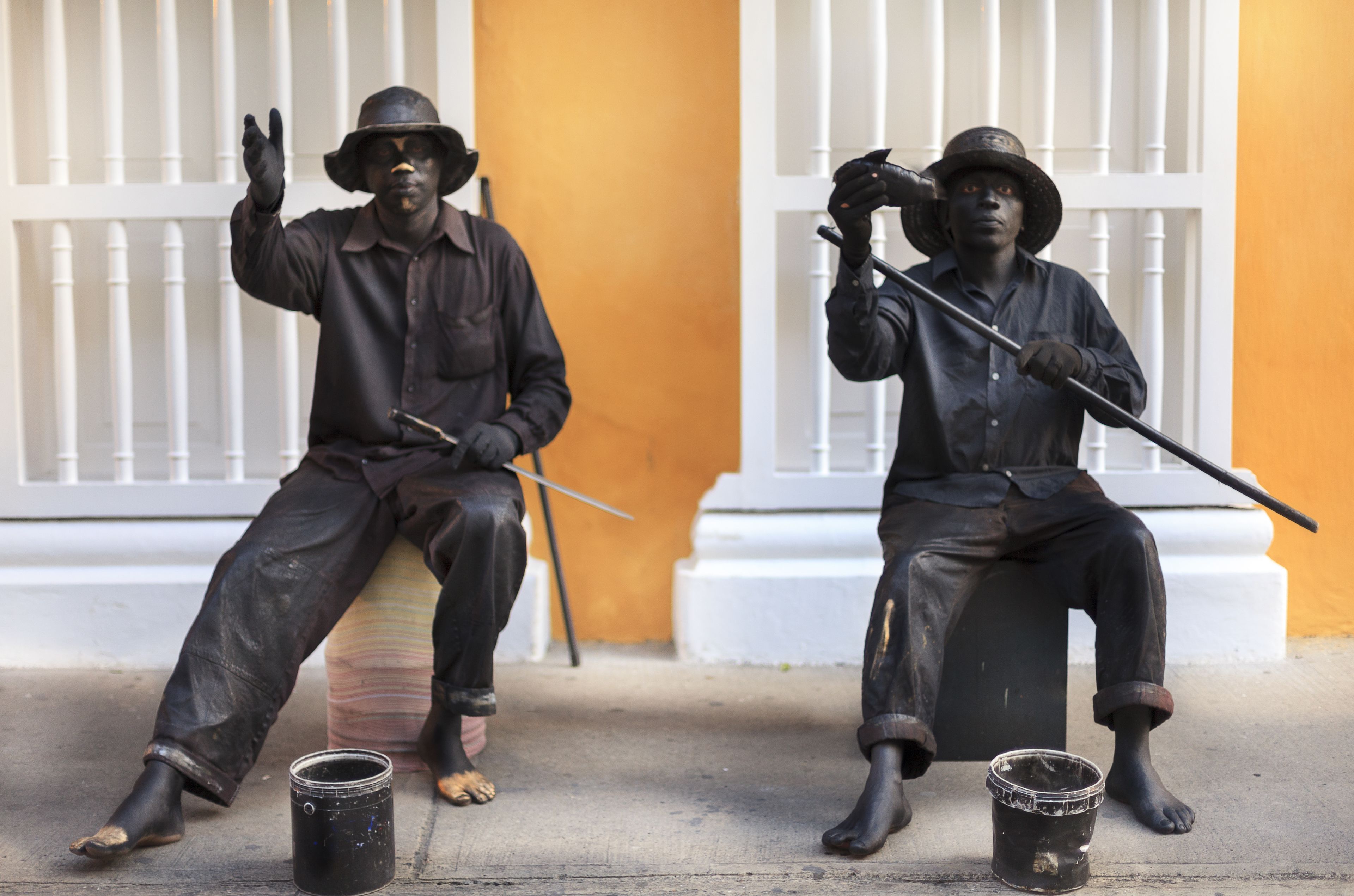 Dos estatuas de hombres sentados en bancos, uno sosteniendo una cámara y el otro un bastón, con cubos a su lado.