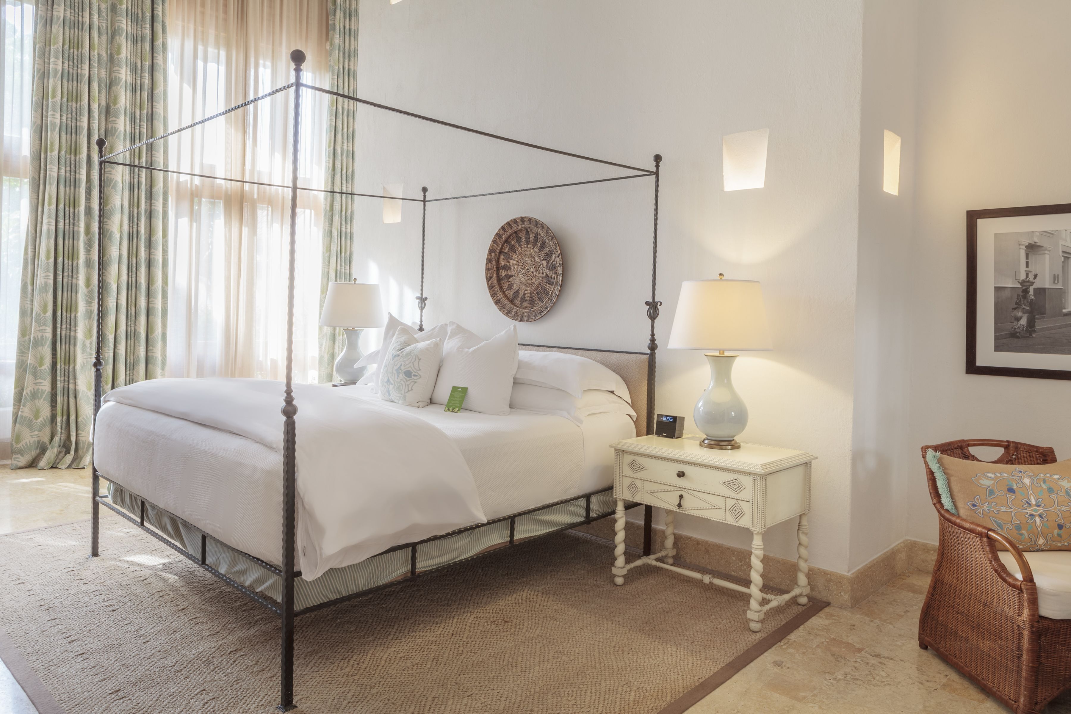 A four-poster bed with a white bedspread, two bedside tables with lamps, a wicker chair, and a wall-mounted round mirror.