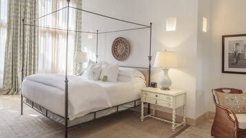 A four-poster bed with a white bedspread, two bedside tables with lamps, a wicker chair, and a wall-mounted round mirror.