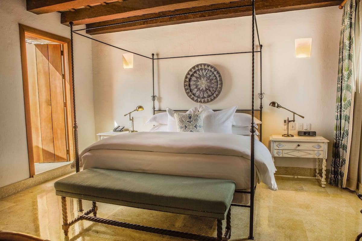 Un letto a baldacchino con una panca di fronte, un comodino con una lampada, un pannello decorativo a parete e una porta in legno.