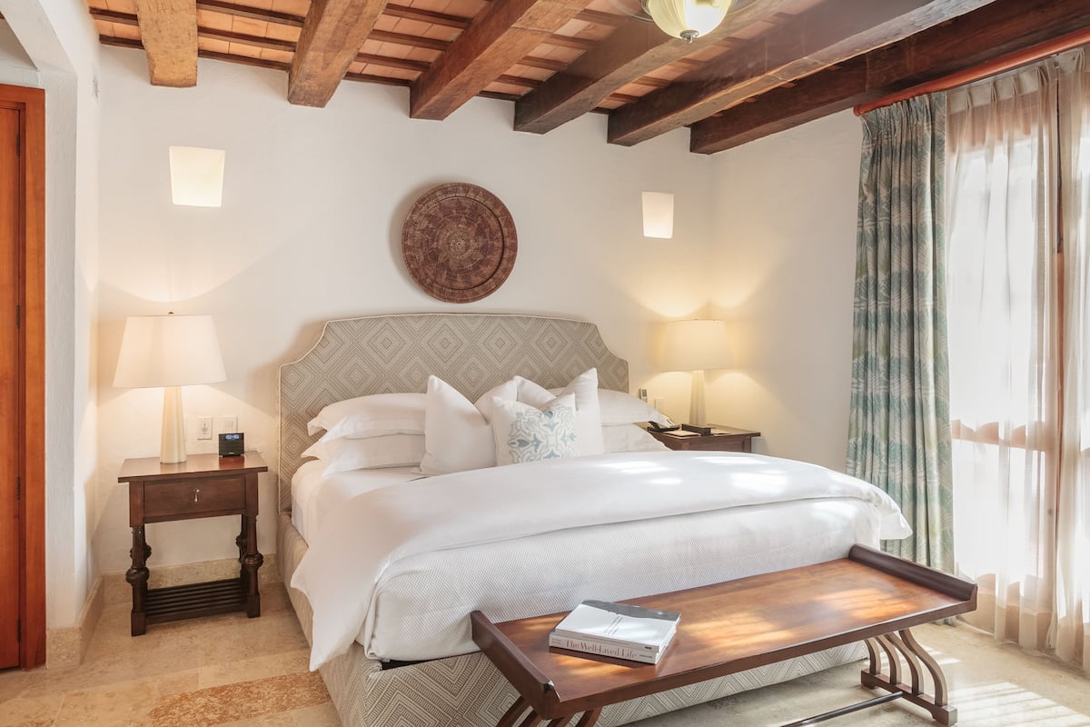 Una camera d'albergo con un letto grande, due comodini con lampade, un tavolino in legno e vista all'esterno attraverso tende leggere.