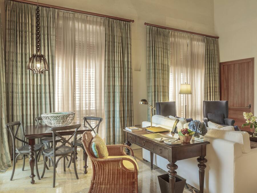 A spacious living room with a dining area, featuring a large wooden table, wicker chairs, a sofa, and a desk with a lamp.