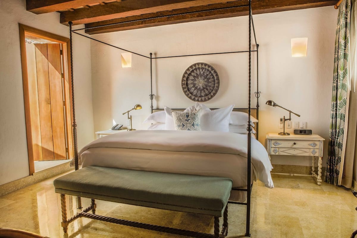 Un letto a baldacchino con una panca, comodini con lampade, una sveglia a parete e un pannello decorativo.