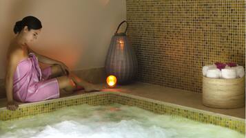 A person sitting by a hot tub with a towel wrapped around their legs, a candle, and a basket of towels.