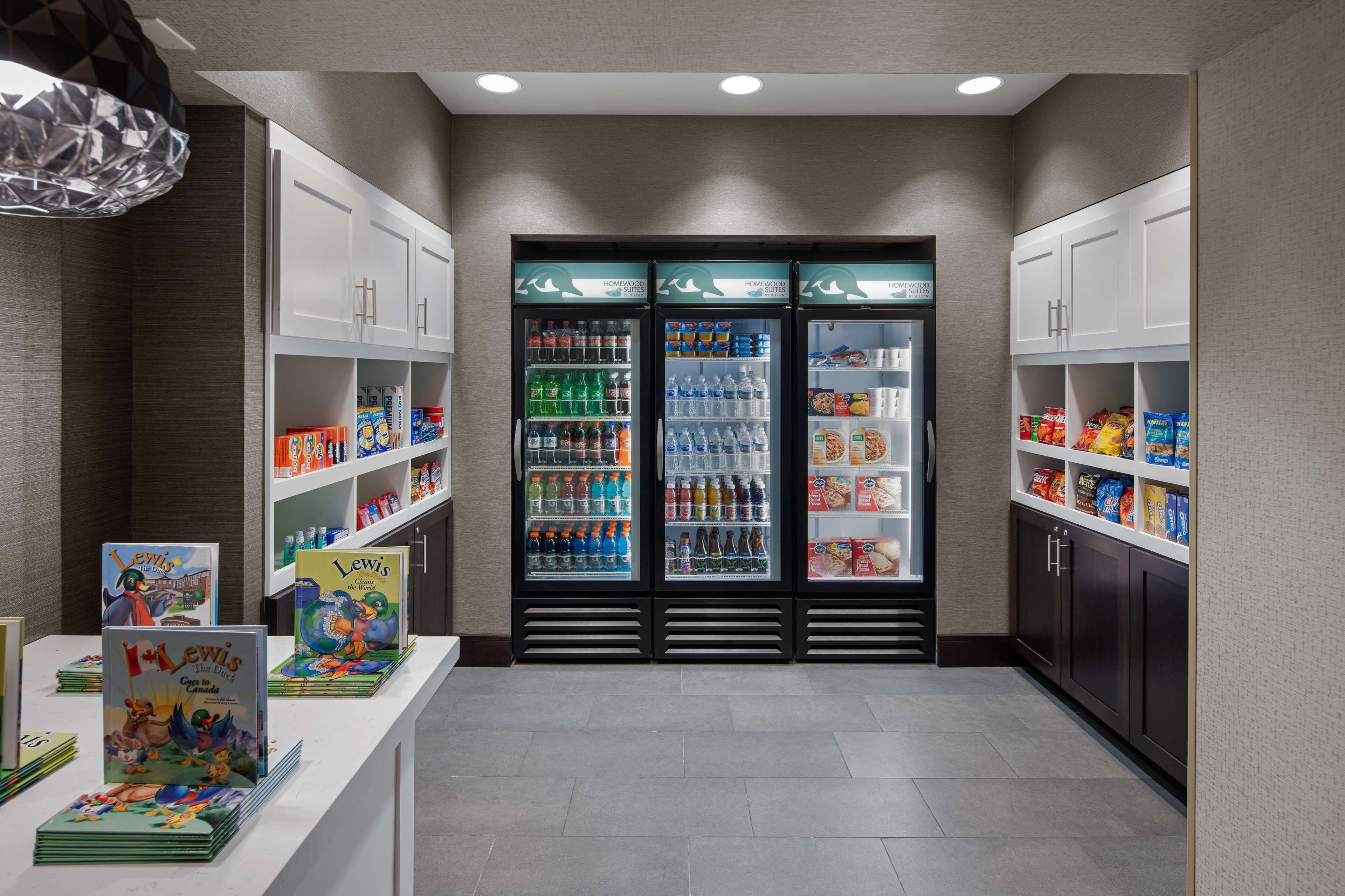 A clean, modern lobby with a beverage refrigerator stocked with various drinks, a children's book display, and neatly organized shelves with snacks and toiletries.