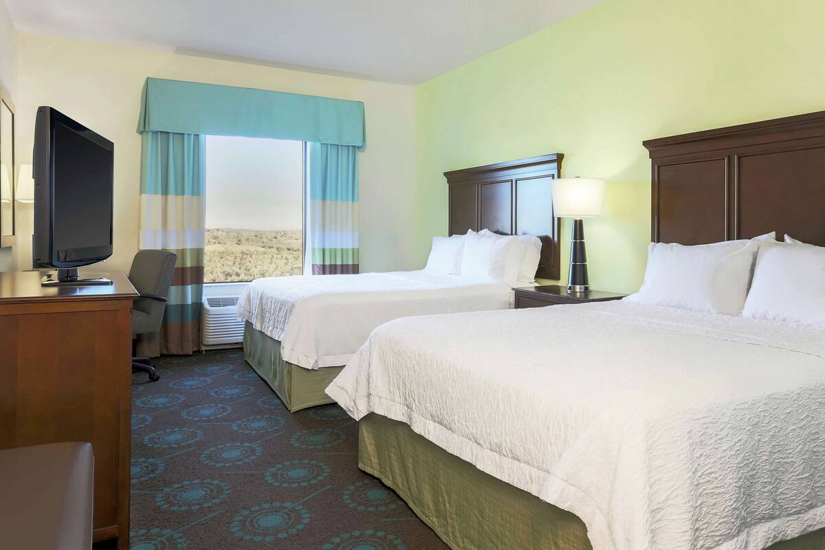 A hotel room with two beds, a desk with a TV, a chair, and a window with curtains.
