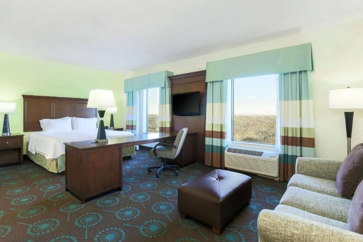 A hotel room with a bed, desk, chair, and ottoman, a TV, and large windows with curtains.
