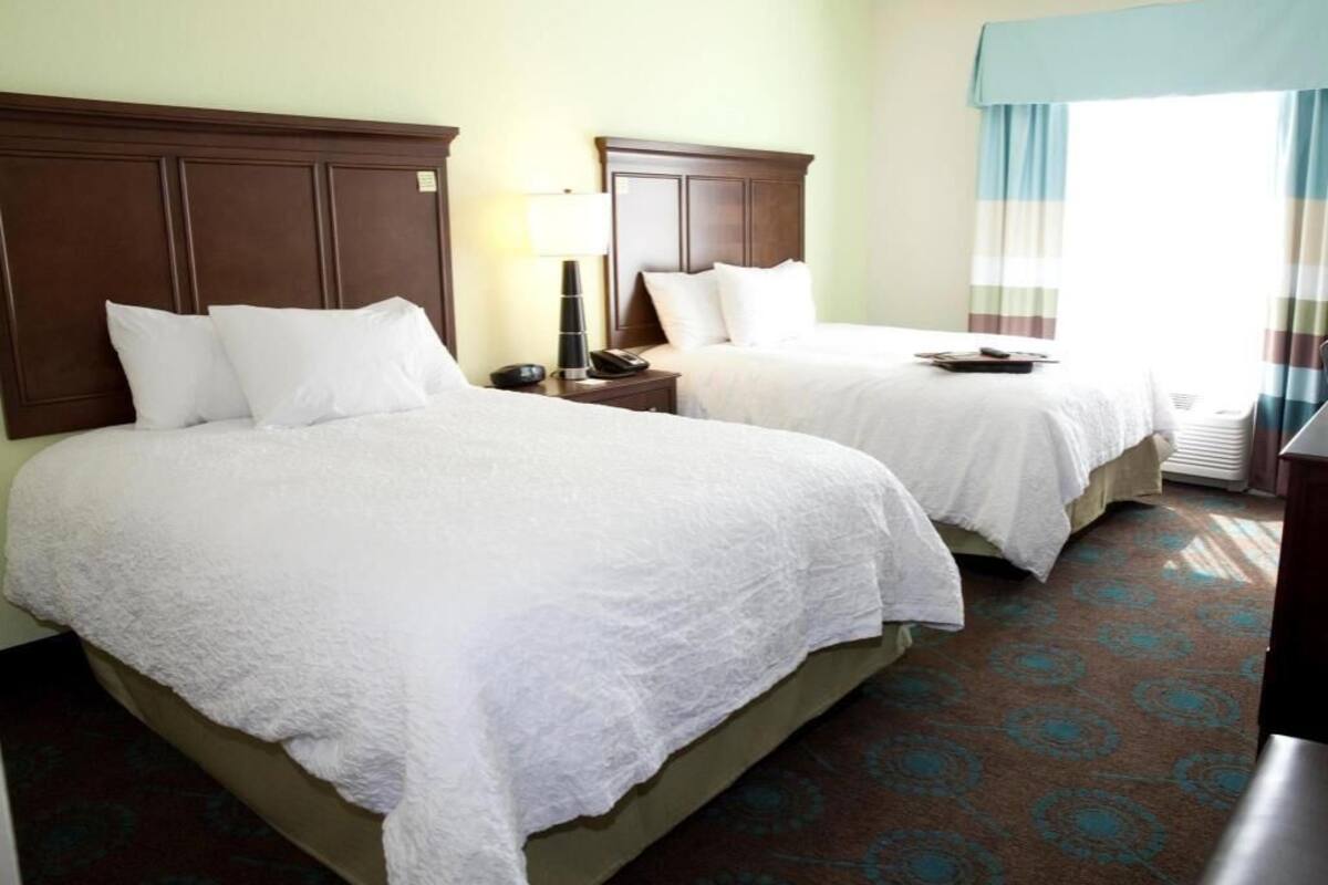 A hotel room with two beds, a nightstand with a lamp, a window with curtains, and a carpet with a circular pattern.