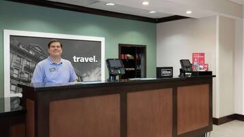 A hotel reception desk with a large travel-themed poster, a digital kiosk, and a minibar cabinet.