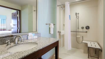A bathroom with a granite countertop sink, a mirror, a shower with a handheld showerhead, and a small stool.