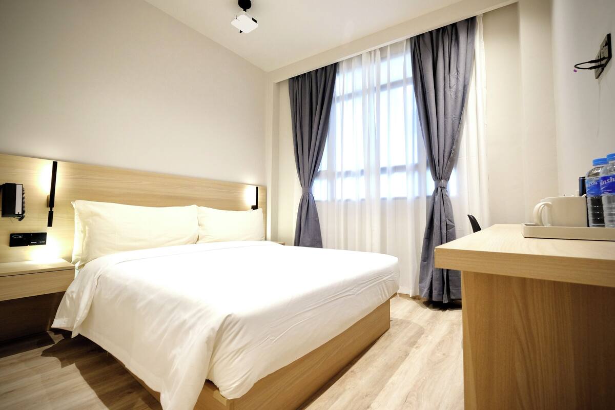 A hotel room with a large bed, bedside tables, a desk with a chair, and a window with curtains.