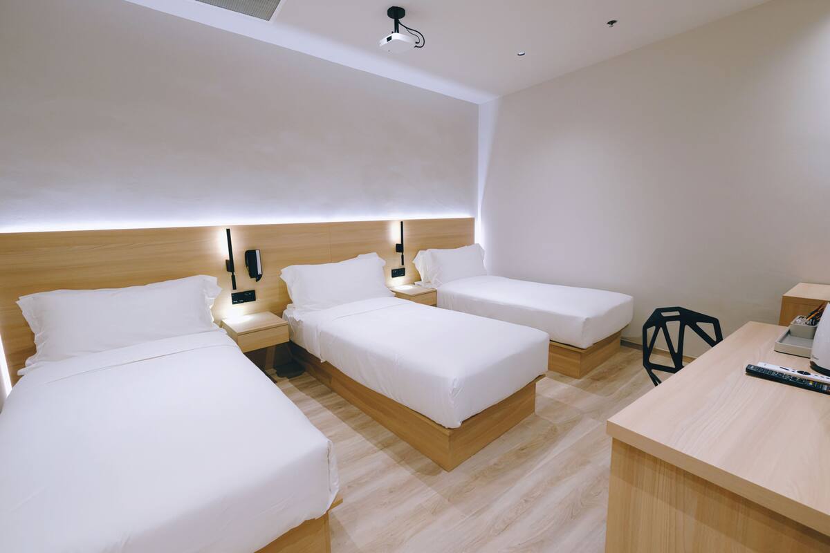 A modern hotel room with three single beds, a desk, and a chair.