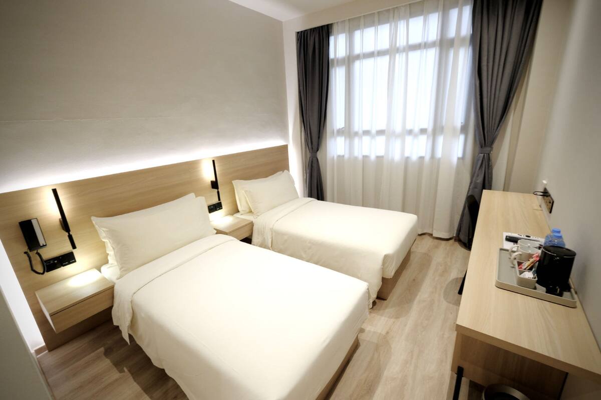 A hotel room with two beds, a desk, a nightstand, and a window with curtains.