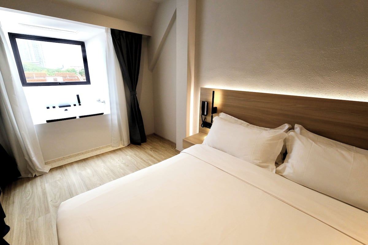 A hotel room with a large bed, a window with curtains, a bedside table with a lamp, and a wooden headboard.