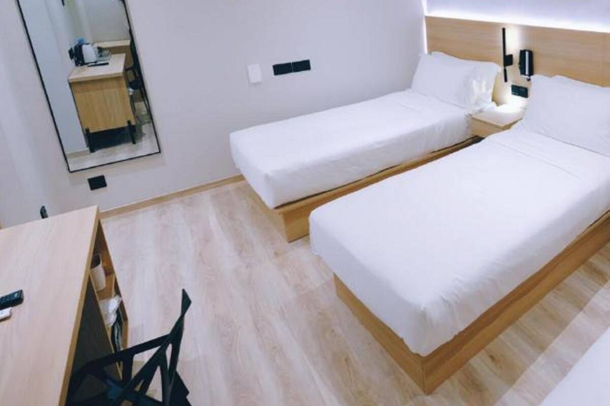 A hotel room with two single beds, a wooden headboard, bedside tables with lamps, a small desk, and a mirror.