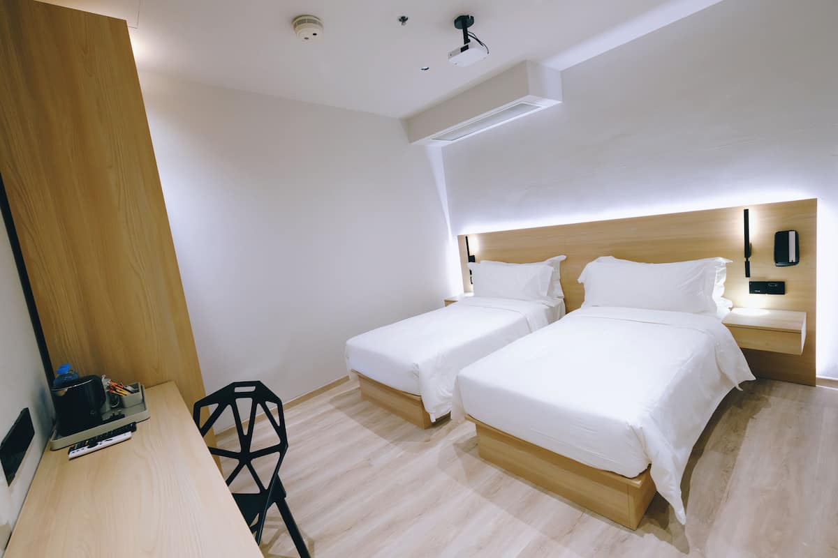 A hotel room with two single beds, a wooden desk, a chair, and a small table with a remote control.