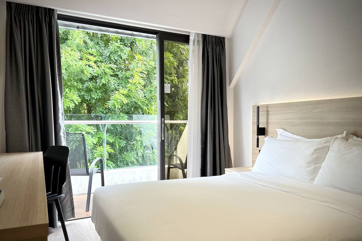 A hotel room with a large bed, a desk, and a chair. There is a window with curtains, and a view of greenery outside.