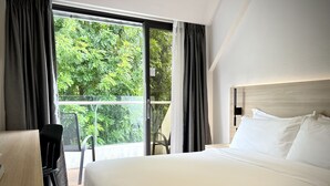 Habitación de hotel con una cama grande, un escritorio y una silla. Tiene una ventana con cortinas, y se ve follaje afuera.